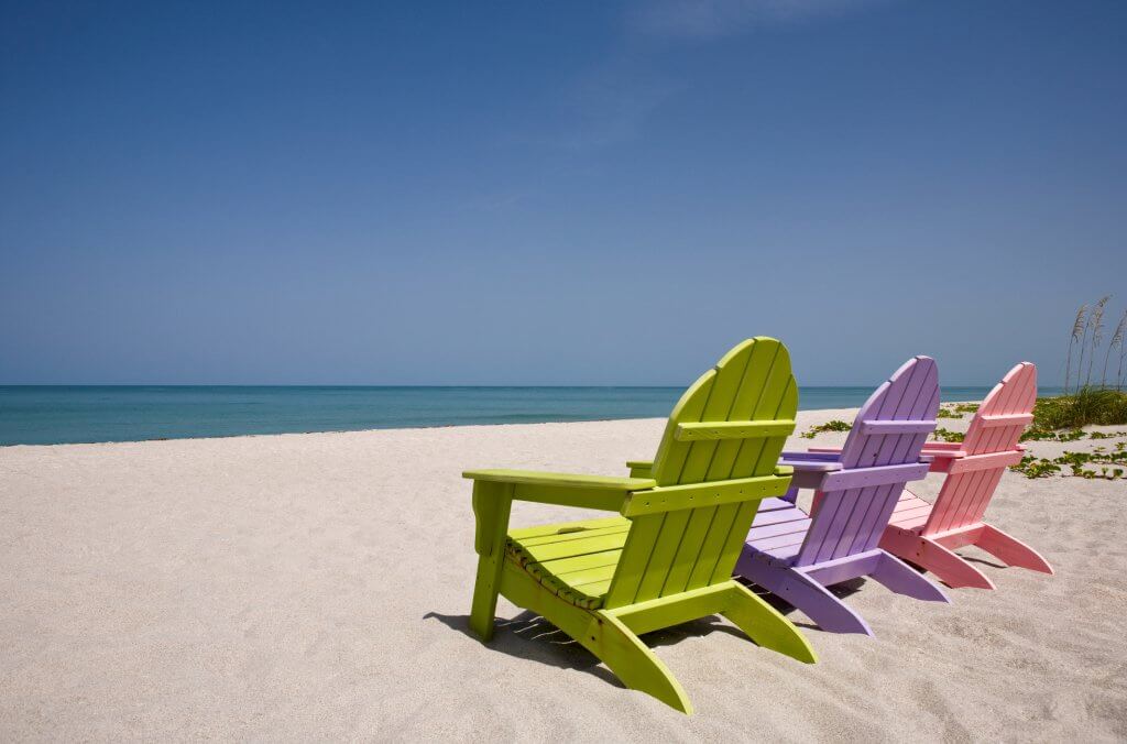 Three pastel colored beach chairs on sand overlooking a turquoise sea. 